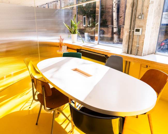 The image shows a brightly lit meeting room with an oval white table surrounded by colorful chairs, against a backdrop of a shiny metallic wall.