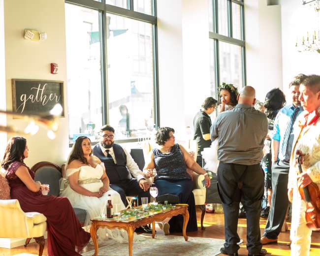 A group of people socialize at a brightly lit venue, with some seated and others standing, while a musician plays the trumpet in the foreground.