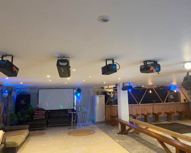 A spacious indoor venue featuring a seating area with a couch, a screen at the front, and a bar setup with colorful lighting and projection equipment.