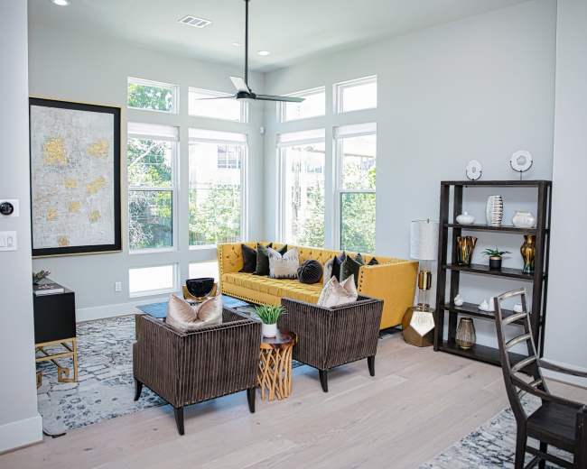 The living room features a yellow sofa, two dark brown armchairs, and a wooden shelf, with large windows allowing natural light to fill the space.