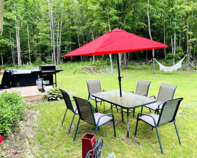 A red umbrella shades a table surrounded by chairs in a grassy backyard near a grill and a hammock.
