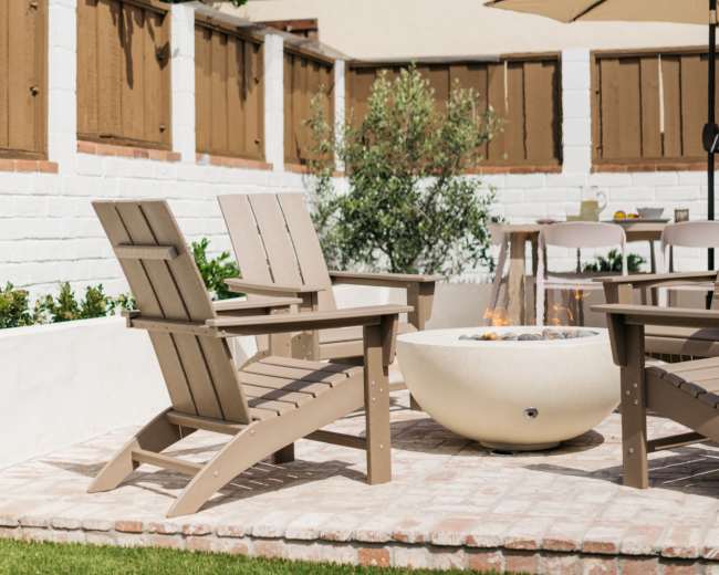 A cozy backyard features wooden Adirondack chairs arranged around a fire pit, with a patio umbrella and greenery surrounding the area.