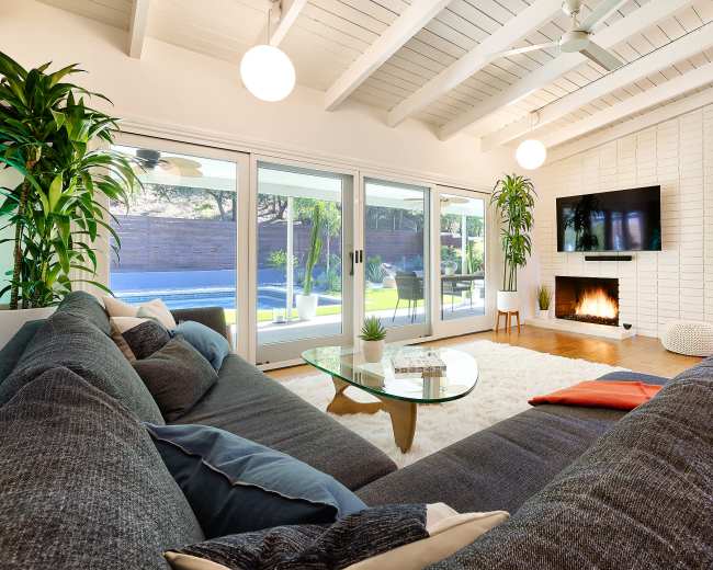 The image shows a modern living room featuring a large sectional sofa, a glass coffee table, a fireplace, and large windows that overlook a backyard with a swimming pool.