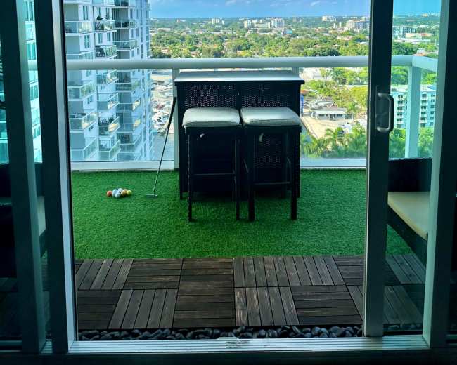 The image shows a balcony with a small table and two chairs, overlooking a cityscape with green trees and buildings under a blue sky.