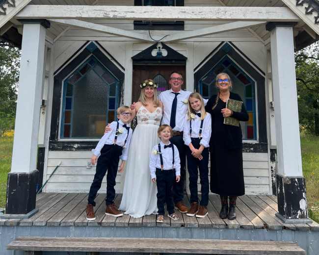 A wedding party poses in front of a vintage house with decorative features and large, colorful windows.
