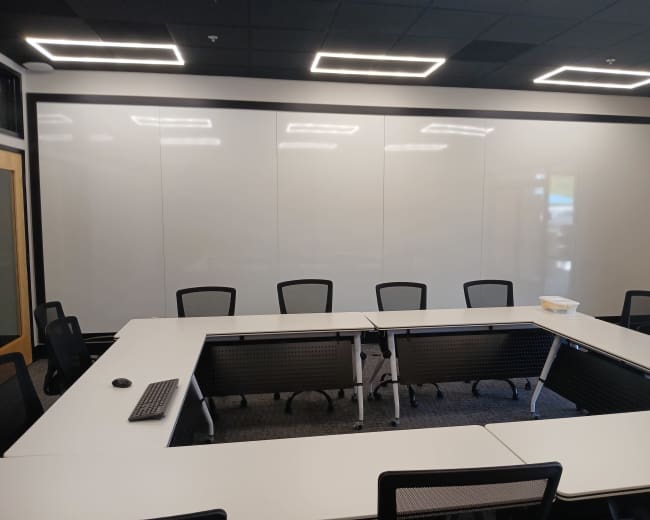 The image shows a modern conference room with a U-shaped arrangement of white tables and black mesh chairs, featuring a large whiteboard wall.