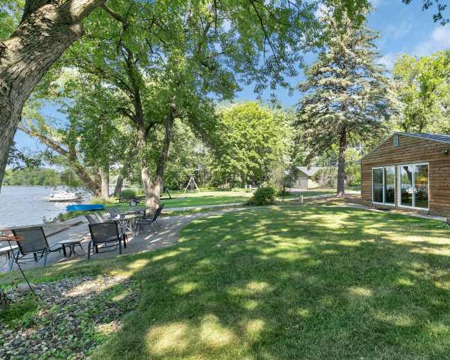 A landscaped area by a lake with a wooden house, trees, and a seating arrangement.