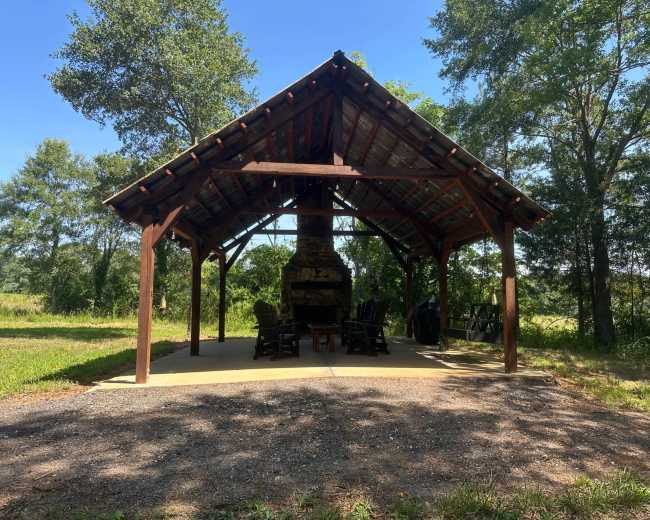 A covered outdoor area with a stone fireplace and several wooden chairs is surrounded by trees and greenery.