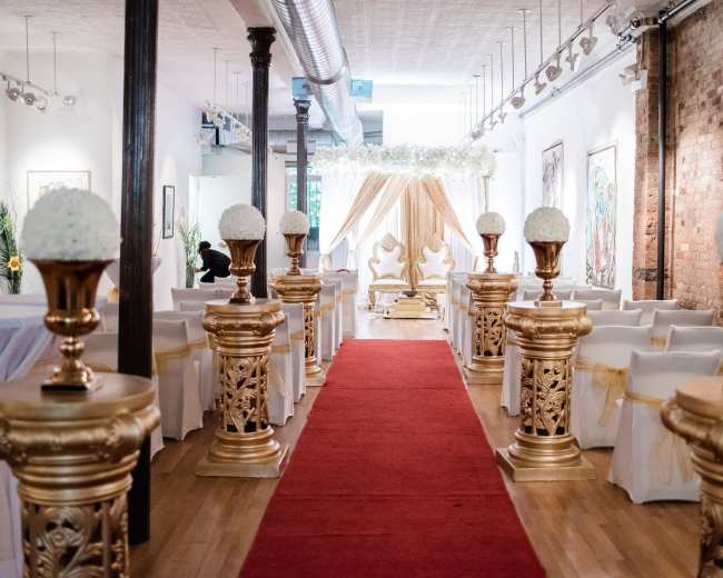 The image shows an elegantly arranged event space with a red carpet leading to a decorated altar, flanked by gold pedestals topped with floral arrangements.