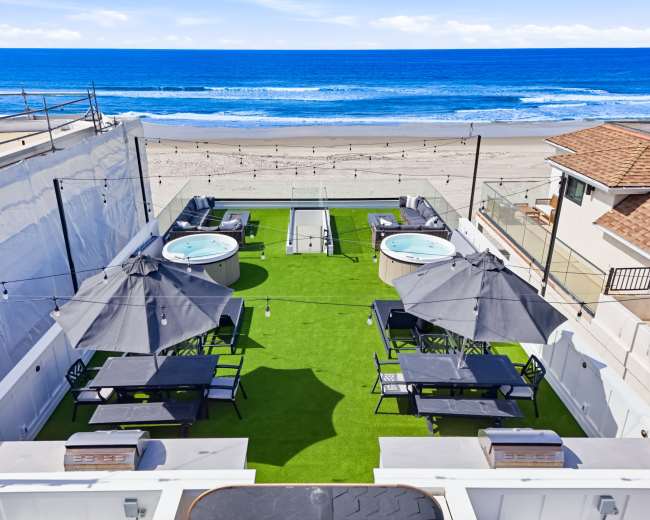 The image shows a rooftop terrace featuring several hot tubs, dining tables, and umbrellas, overlooking a beach and ocean.