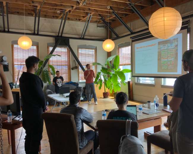 A person is giving a presentation to an audience in a spacious room with plants and large windows.