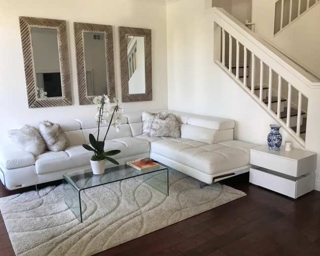 A modern living room with a white sectional sofa, a glass coffee table on a patterned rug, and three mirrors on the wall above.