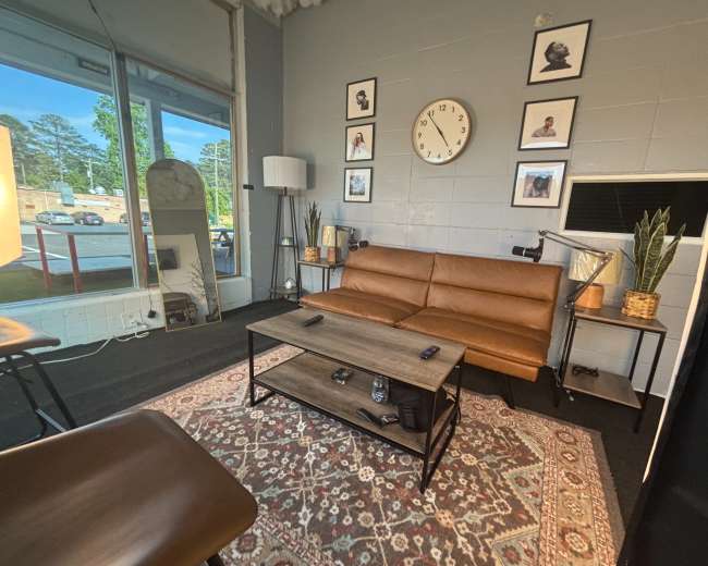The image shows a modern interior space with a brown leather couch, a wooden coffee table, decorative plants, a round wall clock, and framed pictures on the wall.