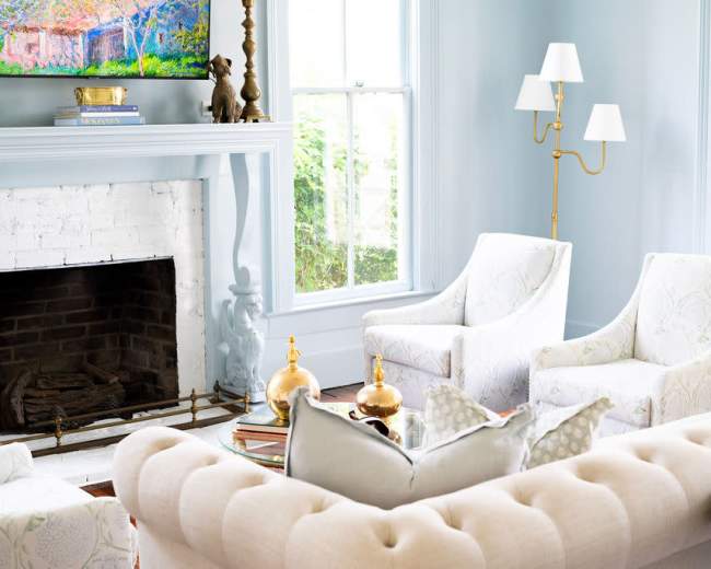 A bright living room features two upholstered armchairs, a beige tufted sofa, and a fireplace with decorative items, all arranged under soft, natural light from large windows.