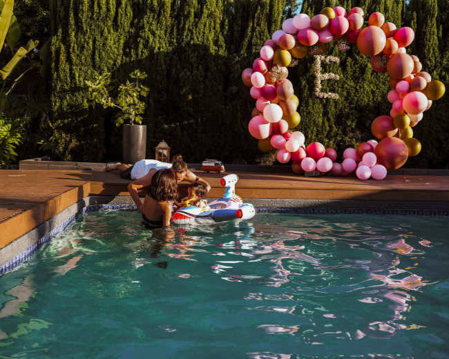 A woman and a child play in a swimming pool surrounded by a wooden deck and a decorative balloon arch.