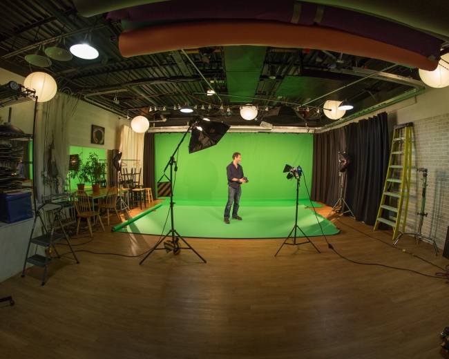 A man stands in a studio with green screens, studio lights, and various equipment arranged around him.