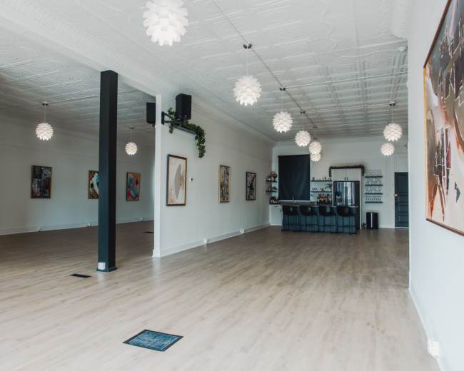 The image shows a spacious, well-lit interior with white walls, wooden flooring, and decorative pendant lights, featuring bar seating and artwork on the walls.