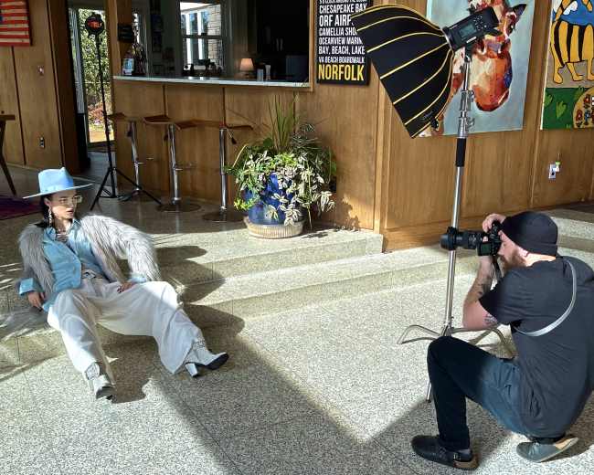 A photographer takes pictures of a model wearing a wide-brimmed hat and a faux fur coat while seated on a floor in a well-lit indoor space.