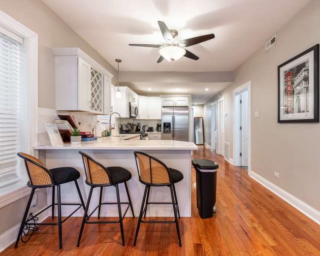 The image shows a modern kitchen with a bar counter featuring three stools, light-colored cabinets, and a doorway leading to a hallway.