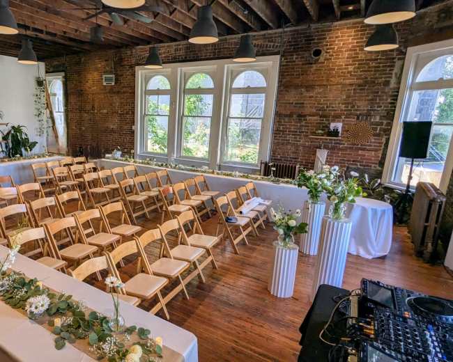 The image shows a spacious indoor venue set up for a wedding, featuring rows of wooden chairs, tables with floral arrangements, and large windows allowing natural light.