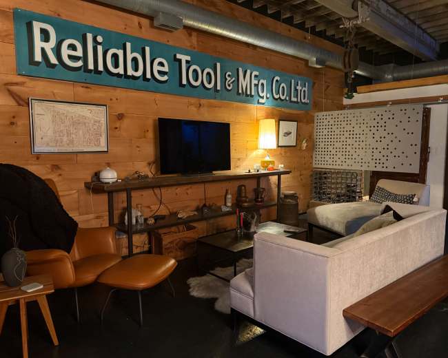 The scene shows a cozy living area with a wooden wall featuring the sign "Reliable Tool & Mfg. Co. Ltd., a television on a shelf, a couch, and a wooden bench.