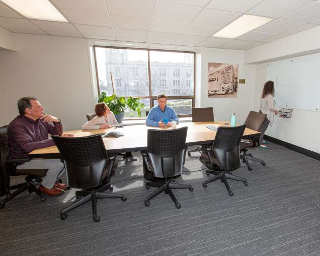 A group of four people is seated at a conference table in a well-lit meeting room, with one person writing on a whiteboard while others observe.