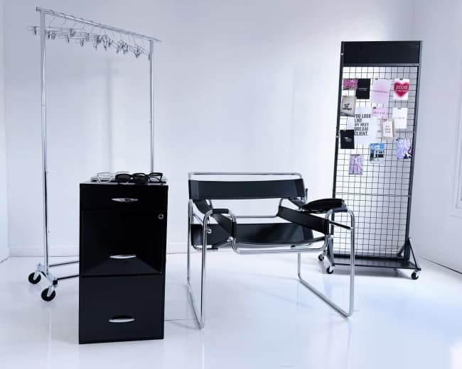 The image shows a minimalist interior featuring a black rolling storage unit, a modern chair, a clothing rack with hangers, and a grid display board with various notes and images.