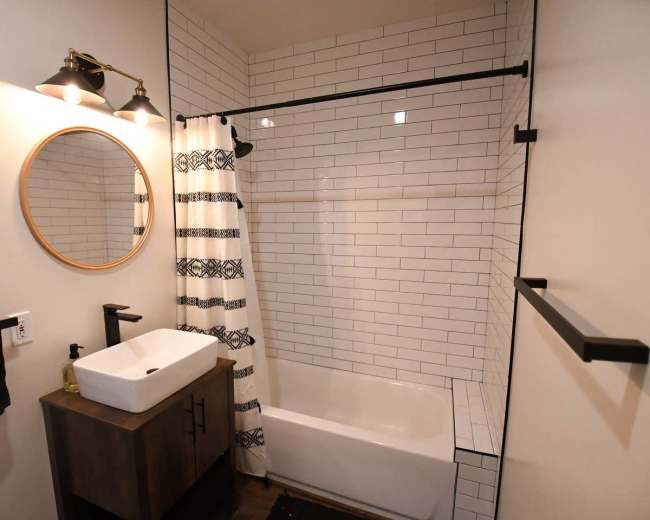 The image shows a modern bathroom featuring a white tiled bathtub with a glass shower rod, a round mirror above a rectangular sink on a dark wood cabinet, and a towel rack with black towels.