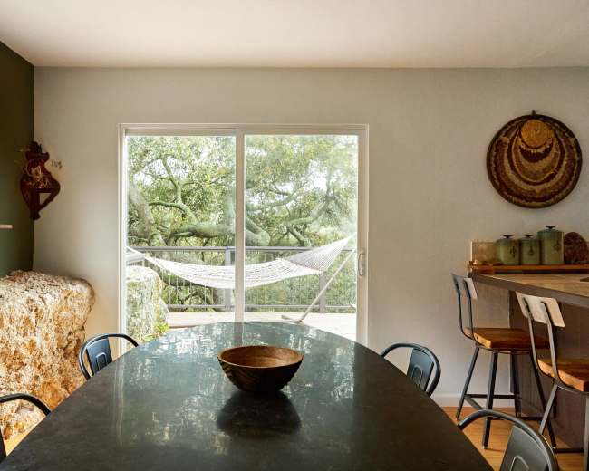 A dining area features a dark table with a wooden bowl, surrounded by chairs, and large windows revealing a view of a hammock outside.