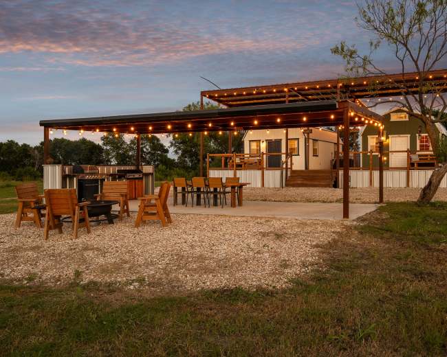 A modern cabin with a covered patio adorned with string lights is set within a grassy area, featuring outdoor seating and dining arrangements.