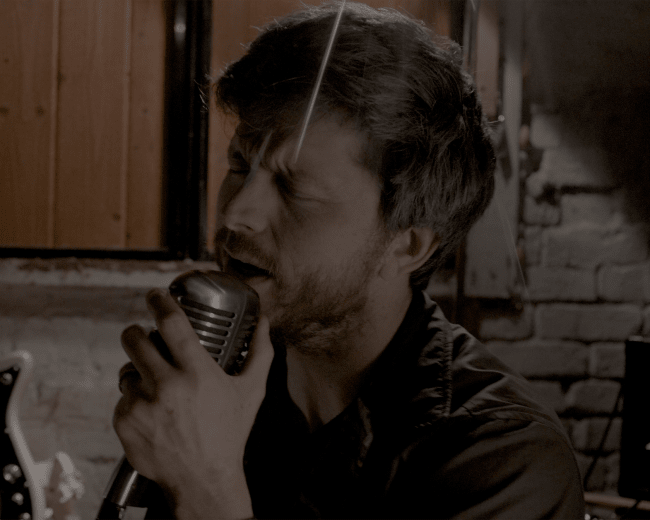 A man sings passionately into a vintage microphone in a dimly lit room with a guitar visible in the background.