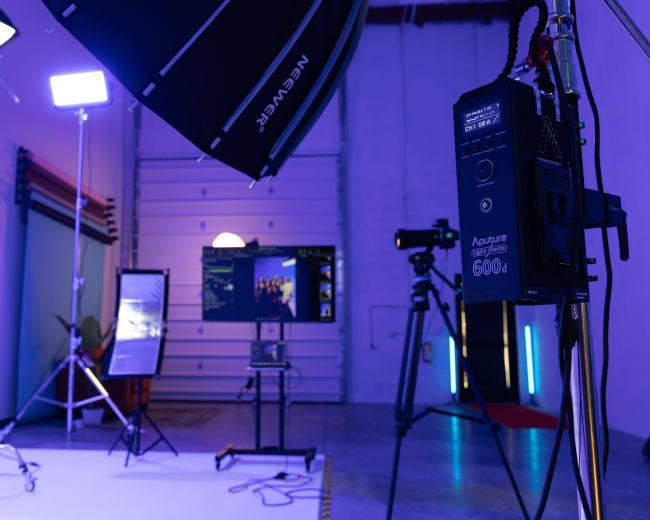 The image shows a photography studio setup with various lighting equipment, a television display, a camera on a tripod, and a backdrop ready for a shoot.