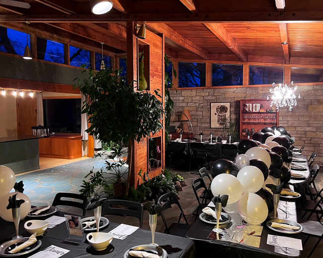 The image shows a decorated indoor gathering space with tables set for a formal event, featuring black and white balloons and a chandelier.