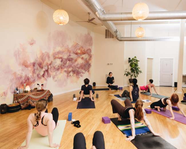 A yoga class is taking place in a studio with participants practicing various postures on mats while a teacher provides guidance at the front.