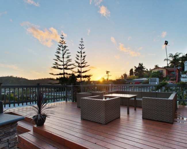 A wooden deck features two chairs and a table, set against a backdrop of a sunset with palm trees and houses in the distance.