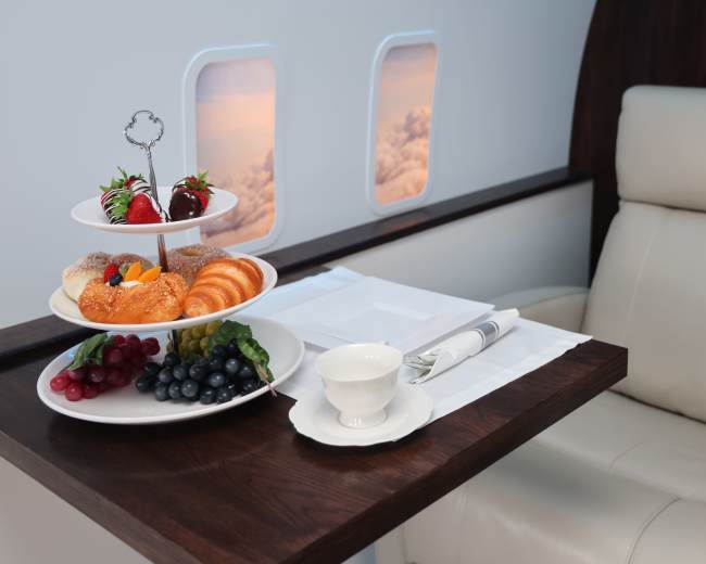 A tiered stand with pastries, fruits, and berries is placed on a wooden table next to a white cup in an airplane seat beside windows showing clouds.