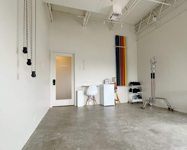 The image shows a minimalist interior space with concrete flooring, a white side table, a chair, and colorful rolls of material leaning against the wall.