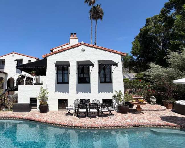 A white stucco house with black window awnings and a red tile roof is situated beside a swimming pool surrounded by lush greenery and decorative plants.