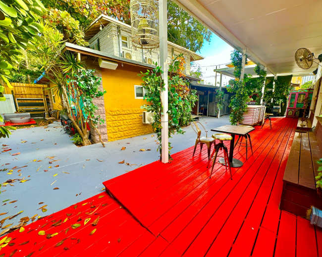 The image shows a vibrant courtyard with a red wooden deck, surrounded by greenery and buildings in the background.