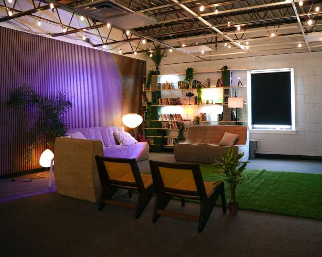 The image shows a cozy lounge area with two sofas, wooden chairs, plants, bookshelves, and ambient lighting.