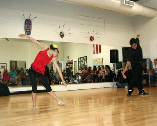 A dancer in a red top performs gracefully while another dancer in black watches in a studio filled with an audience.