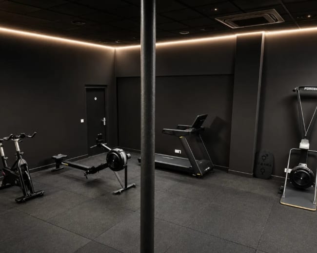 The image shows a dark-themed gym featuring cardio equipment including a treadmill and rowing machine, along with exercise bikes and a central support column.