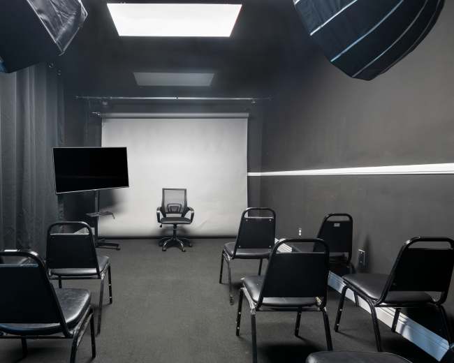 A small, dimly lit room features a center chair facing a blank screen, surrounded by several black chairs and equipped with lighting setups.