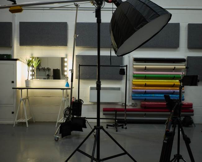 The image shows a photography studio featuring a light setup, a mirror with bulbs, and a backdrop rack with colorful roll paper.