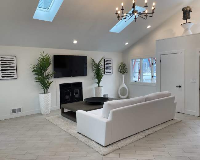 A modern living room with a white sectional couch, a dark coffee table, potted palms, and a television mounted on the wall beneath a skylight.