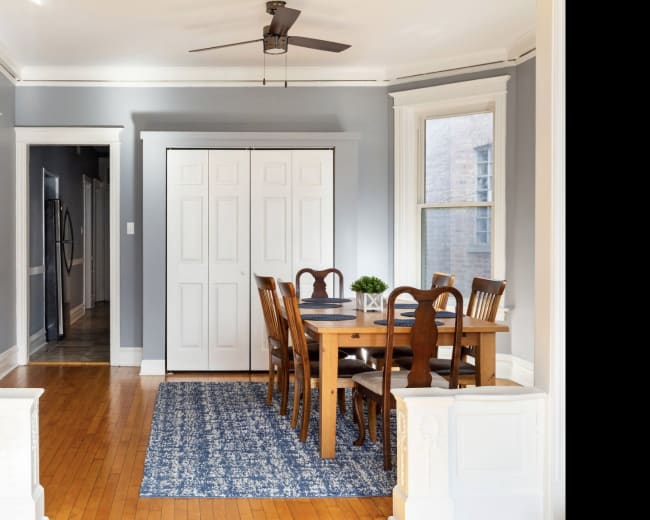 A dining area with a wooden table and chairs, situated on a blue rug, adjacent to light gray walls and a ceiling fan.