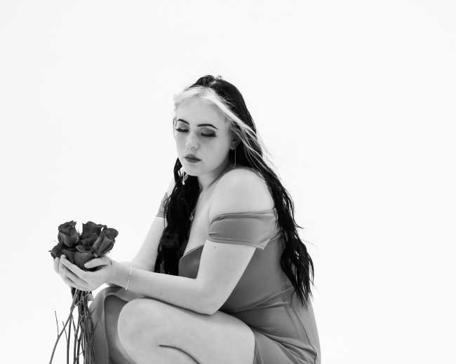 A woman in a long, flowing dress squats on the floor while holding a bouquet of roses.