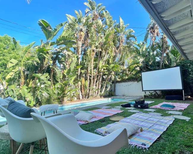 A backyard setup features white chairs and colorful blankets around a projector screen beside a swimming pool, surrounded by lush green palm trees.