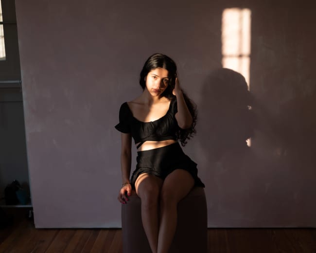 A person sits on a stool in a softly lit room, with a circular mirror framing the scene and casting shadows on the wall.