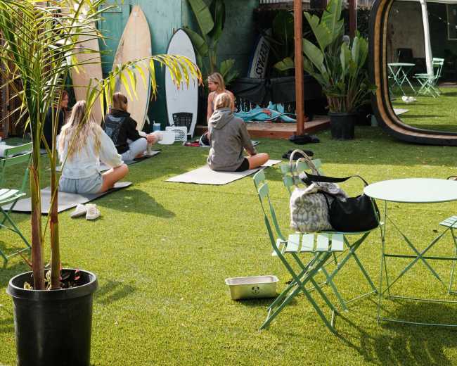 A group of people sits on artificial grass in a communal outdoor space with surfboards and greenery surrounding them.